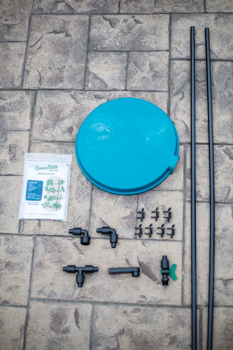 Components of the GreenStalk Watering System in the Dragonfly color are arranged on stone tiles, featuring a blue circular lid, two black poles, an instruction booklet, and a variety of black plastic connectors and clips—everything you need to set up your own automatic watering system.