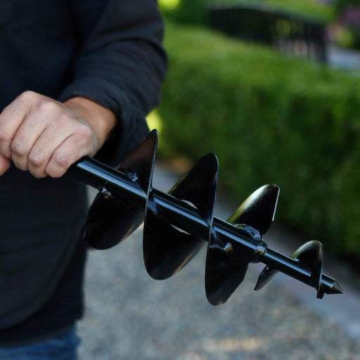 Person holding a black garden auger drill bit with a blurred green background