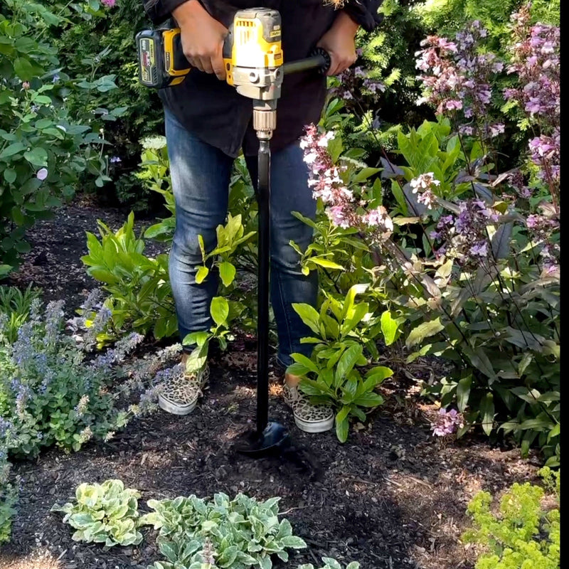 Person using a power tool in a garden setting with plants and flowers.