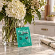 A box of FloraLife Flower Food (Pack of 10), a trusted vase water conditioner for longer-lasting blooms, sits on a marble countertop next to a vase of white and green flowers in a bright kitchen.