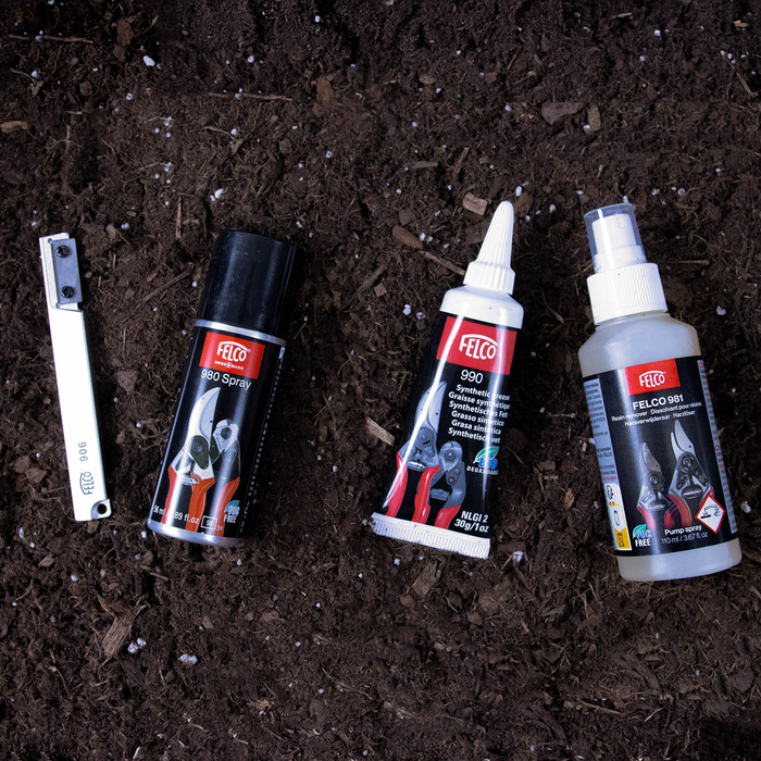 The Felco Maintenance Kit, featuring a metal tool, spray can, lubricant tube, and spray bottle—all clearly labeled for garden tool care—is displayed on dark soil. Designed for comprehensive pruning tool upkeep, this kit is ideal for anyone dedicated to maintaining their Felco pruners and seeking a complete maintenance solution.