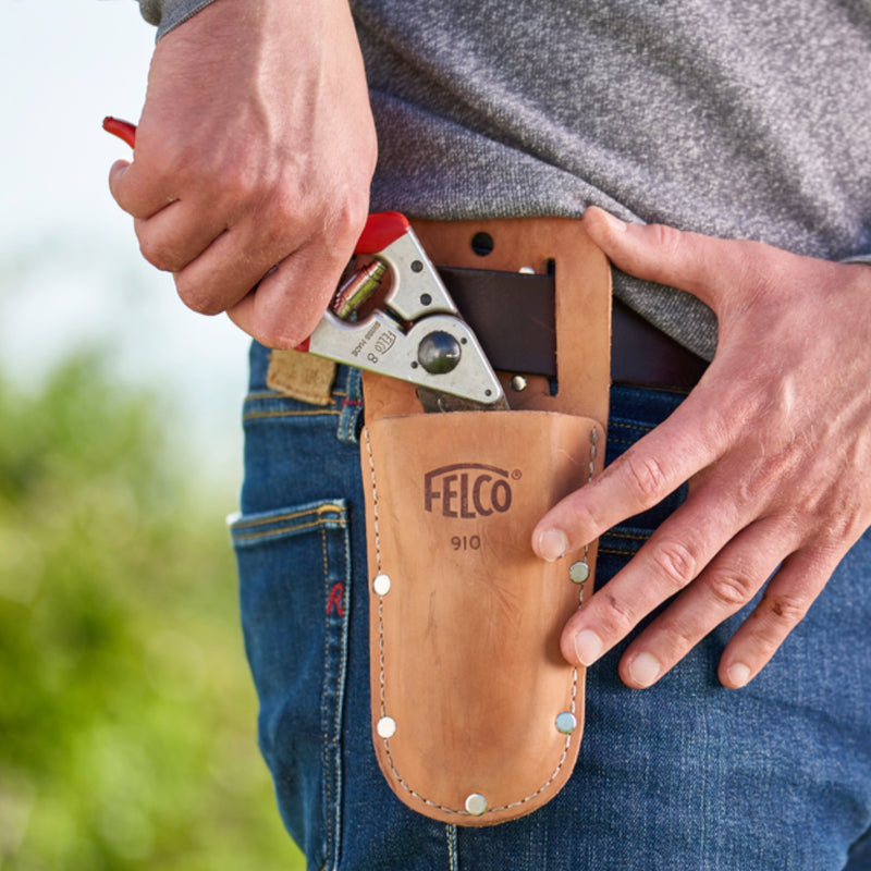 A person wearing a beige Felco 910 leather holster on their belt, holding a pruning shear inside it.