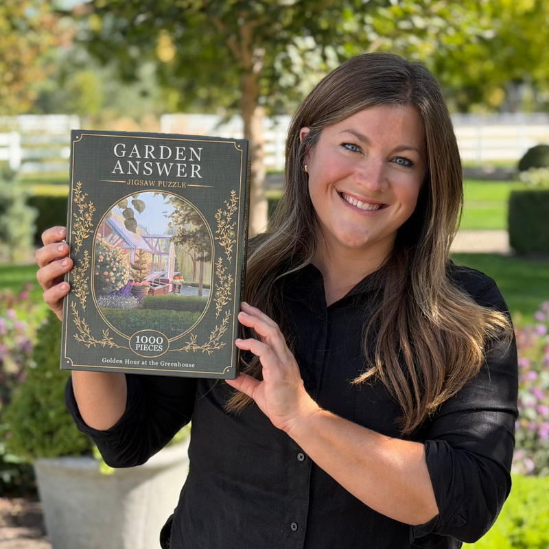 A smiling woman in a black shirt holds up the Golden Hour at the Greenhouse Puzzle (1000 Piece), featuring an illustrated greenhouse scene. She is outdoors with a blurred garden background.
