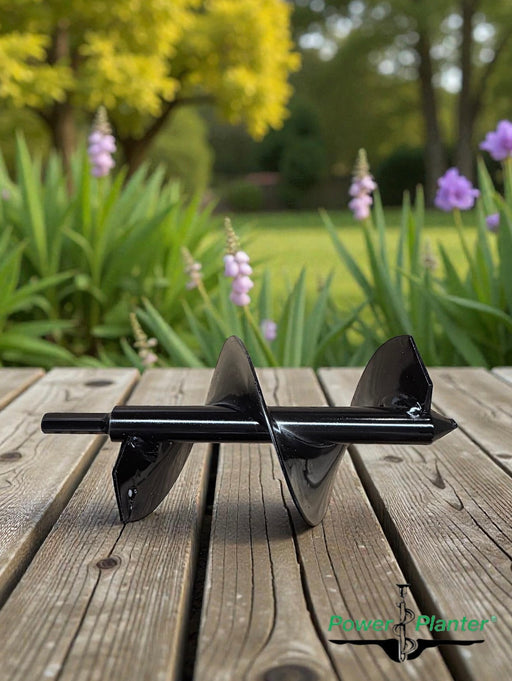 A Power Planter Bulb Planting Auger & Bedding Plant Tool (3″ x 7″), featuring a sleek black design, rests on a wooden surface surrounded by green plants, purple flowers, and trees in the softly blurred background. The Power Planter logo is visible in the bottom right corner.