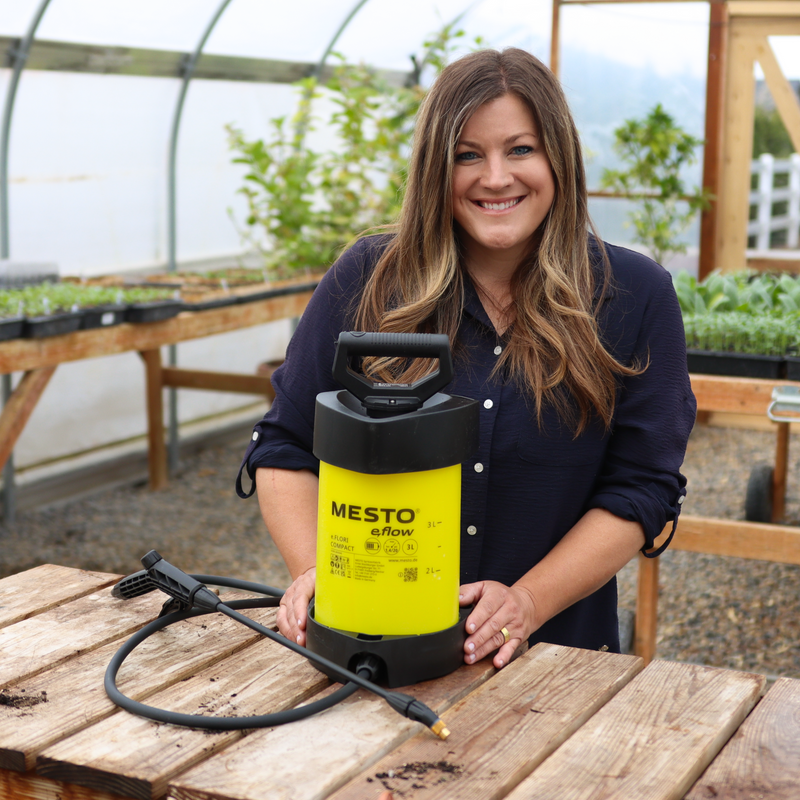 A woman smiles in a greenhouse while standing behind a Mesto E-Flow electric sprayer, yellow and black in color, resting on a wooden table surrounded by soil and plants.
