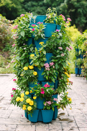 The GreenStalk 7 Tier Leaf Vertical Planter in Dragonfly—the striking basket weave texture version—stands tall on a paved garden path, its multiple tiers overflowing with blooming pink, yellow, and white flowers, creating a stunning example of containerized gardening in a lush oasis of greenery.