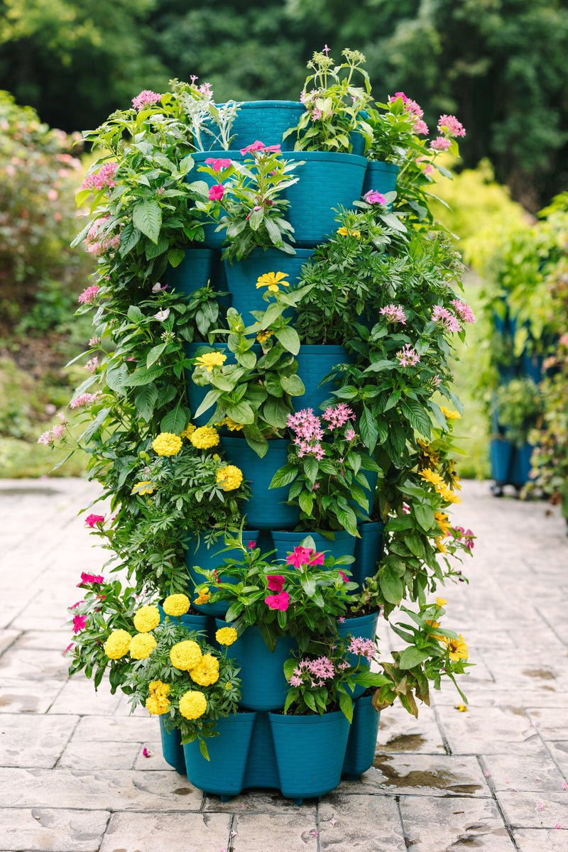 The GreenStalk 7 Tier Leaf Vertical Planter in Dragonfly—the striking basket weave texture version—stands tall on a paved garden path, its multiple tiers overflowing with blooming pink, yellow, and white flowers, creating a stunning example of containerized gardening in a lush oasis of greenery.