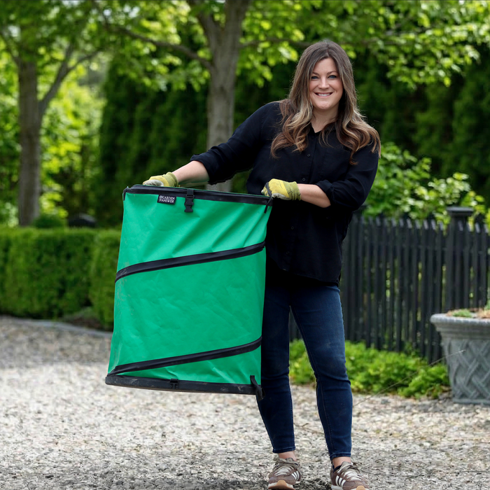 A smiling woman wearing gloves holds the Garden Answer Pop-Up Bag outdoors, surrounded by trees and garden plants on a gravel path.